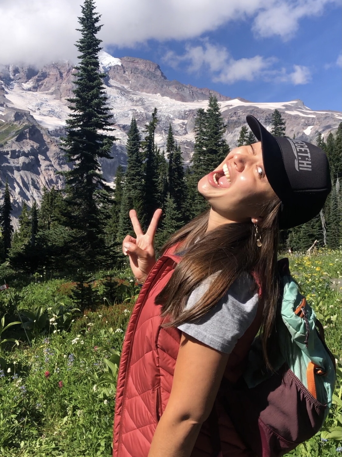 Annalise, a young woman with long brown hair, is winking and smiling at the camera, and making a peace sign with her hands. She is outdoors in front of some pine trees and a snow-covered mountain. 