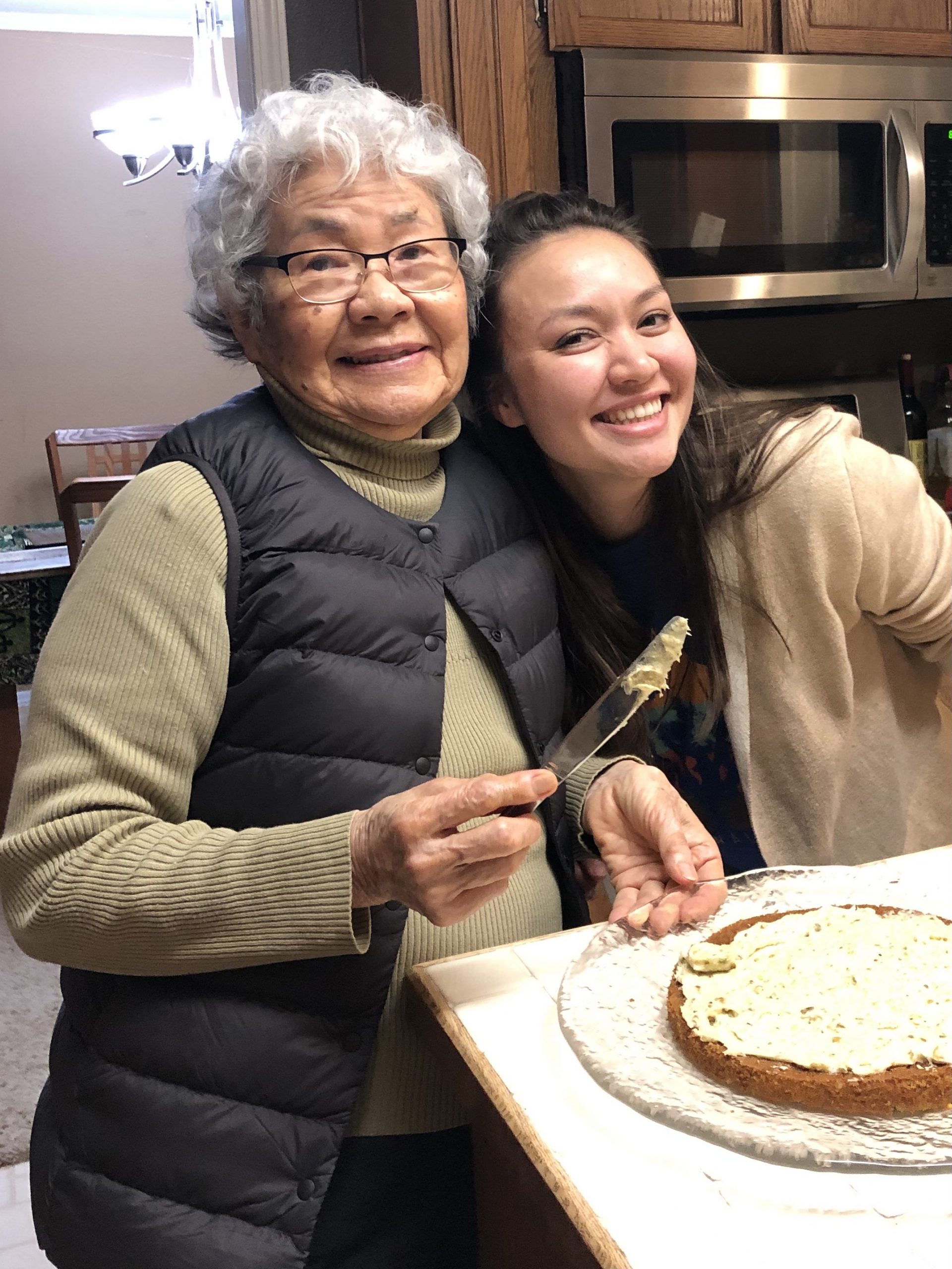 Annalise poses with her grandmother, an Asian woman with short, curly grey hair and glasses. They are making a cake together. 