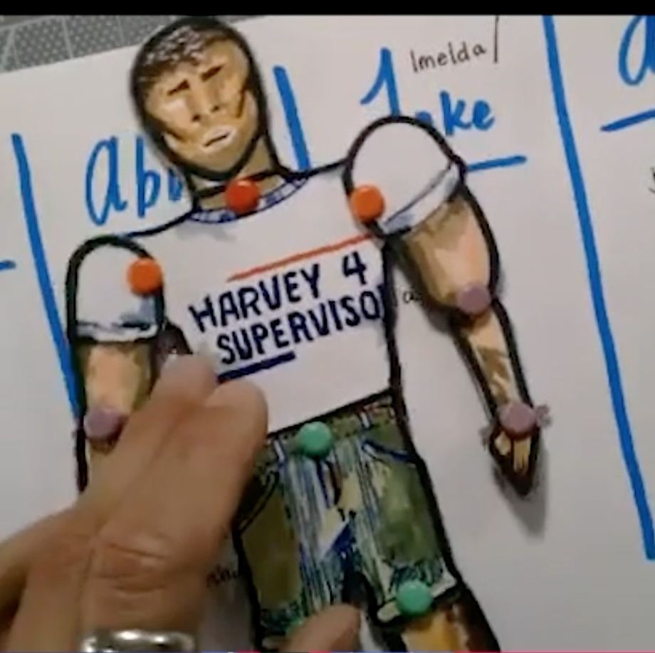 A close-up image of a puppet made from paper and brass brads. The puppet is wearing a shirt that says "Harvey 4 Supervisor"