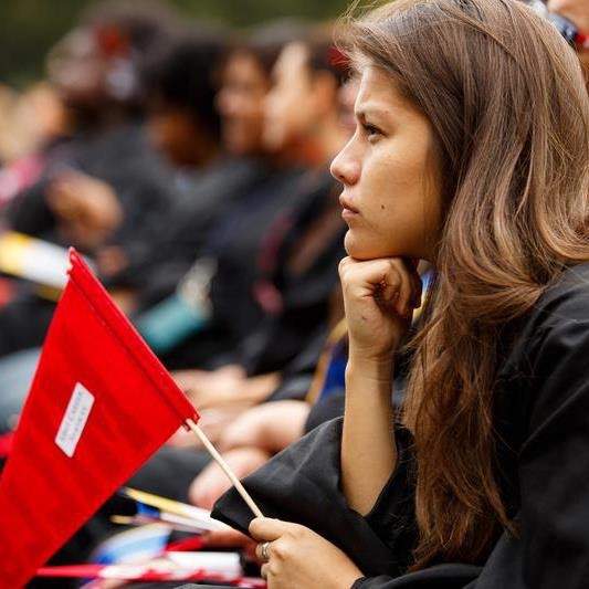 A photograph of Jackie Macy in profile - a Latina woman with long wavy hair wearing a black graduation gown and holding a red flag. She looks pensively away from the camera.